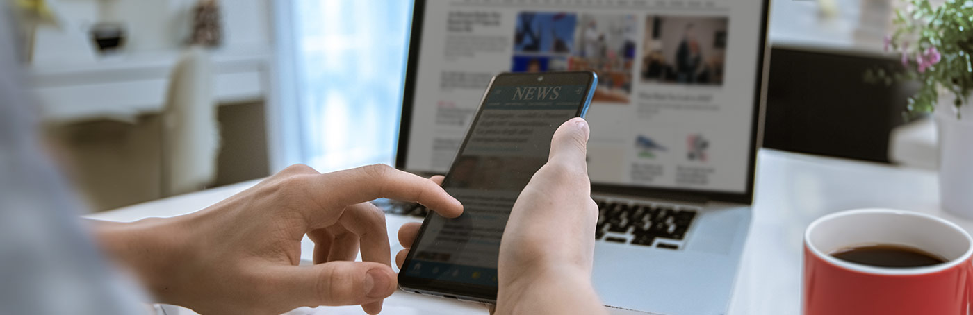 Man reads news article on his phone with a cup of coffee beside him and a laptop in front of him at a table